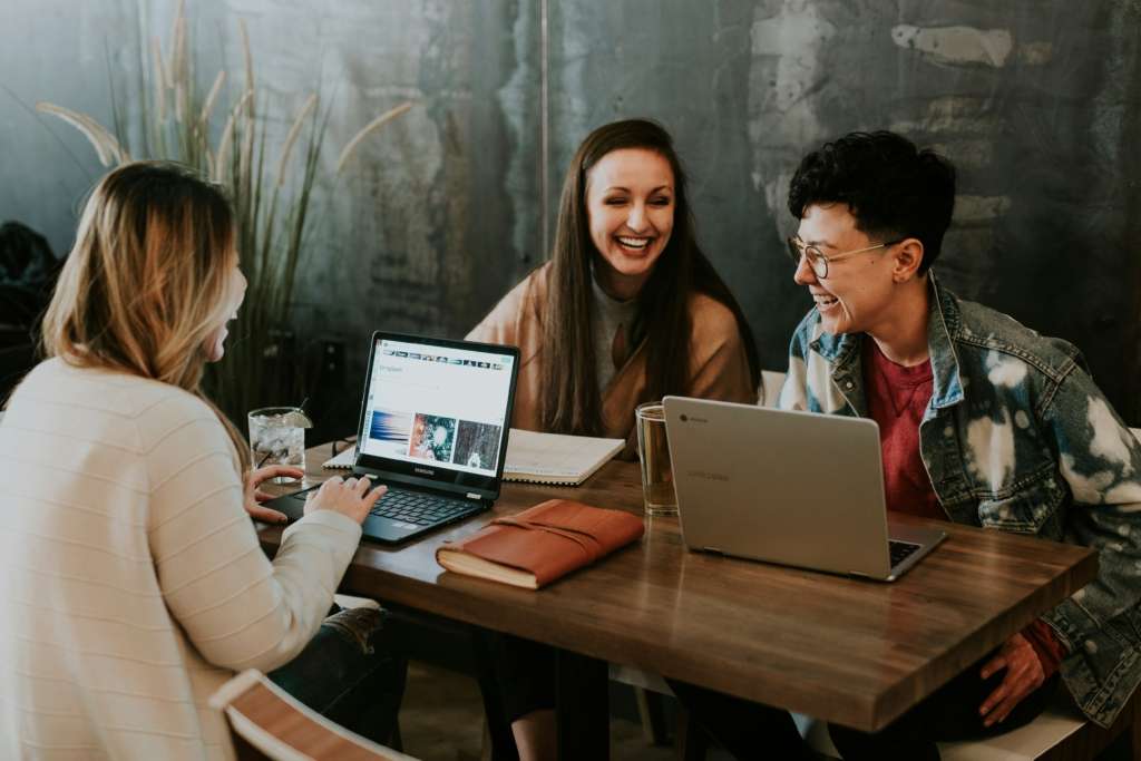 three Gen Z people sitting at a table with laptops laughing together